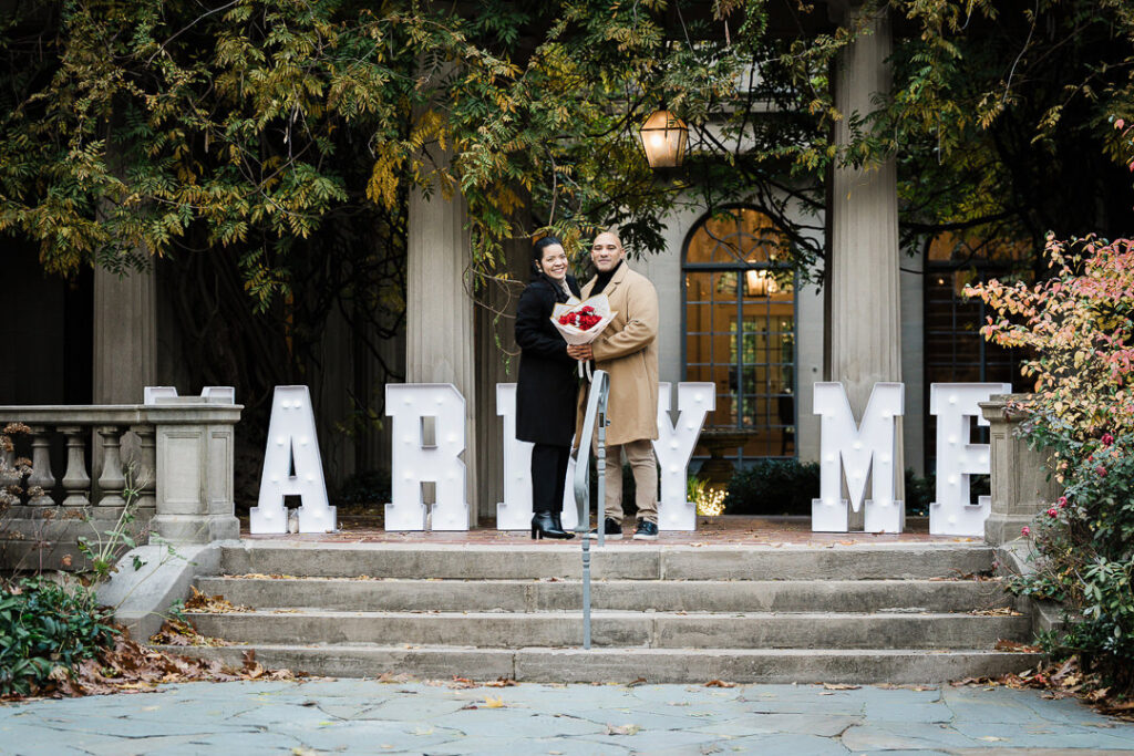 Newly engaged couple poses with red roses and illuminated MARRY ME letters at Van Vleck House & Gardens, professional engagement photography Montclair NJ