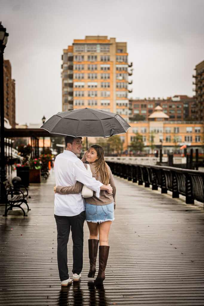 Newly engaged couple under umbrella on rainy Pier 13 in Hoboken with waterfront buildings in background
