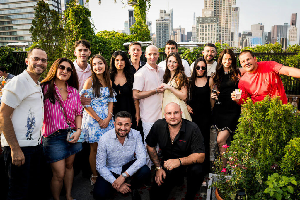 Newly engaged couple celebrates with friends and family in group photo on NYC rooftop with Manhattan skyline, Northern New Jersey proposal photography