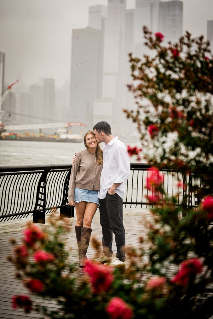 Newly engaged couple walking on Pier 13 with red flowers in foreground and foggy Manhattan skyline behind