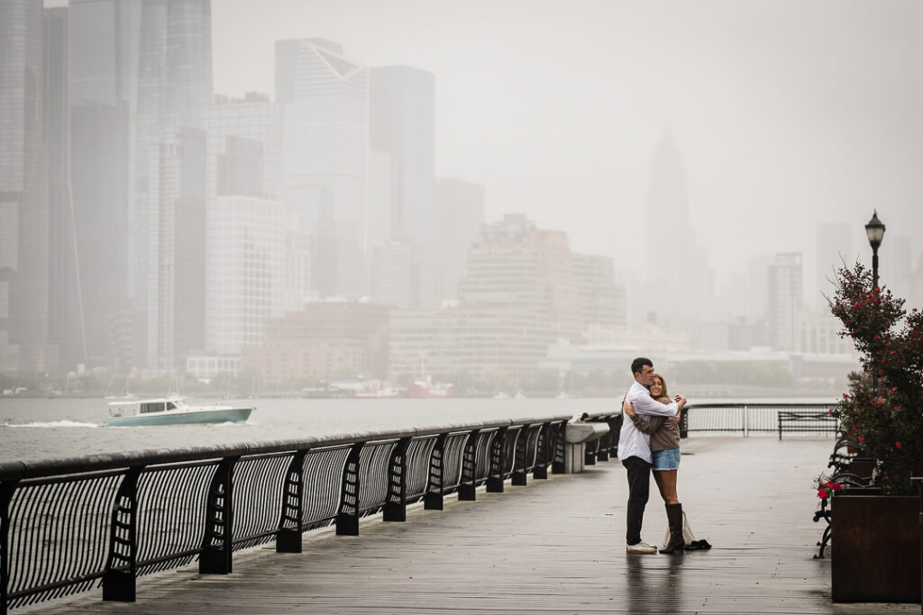 Newly engaged couple standing together on Pier 13 with misty Manhattan skyline and Hudson River in background