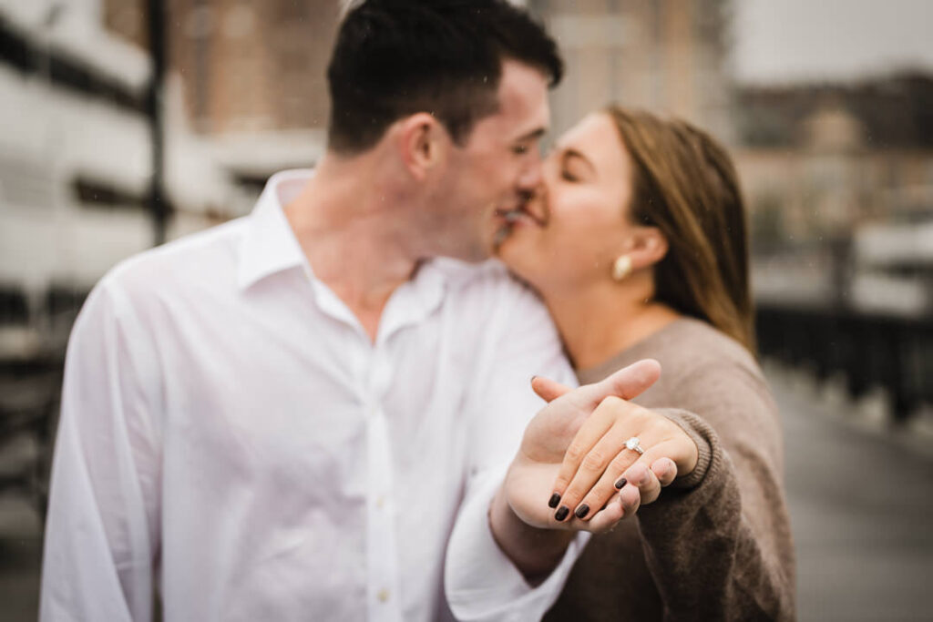 Close up of engagement ring on bride's hand with couple kissing in background after Pier 13 Hoboken proposal