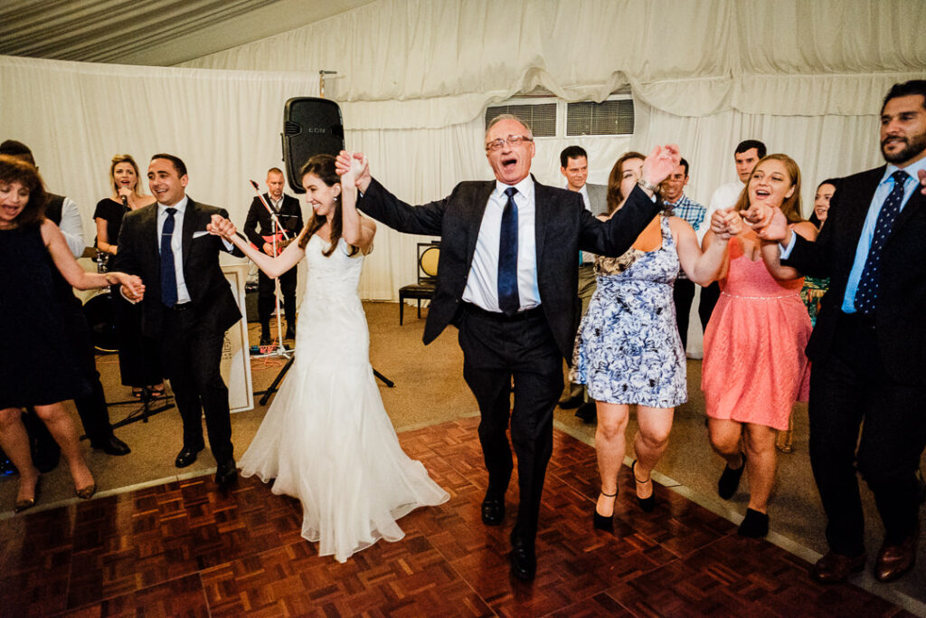 Bride dancing with father during traditional Hora as he is lifted on chair by celebrating guests