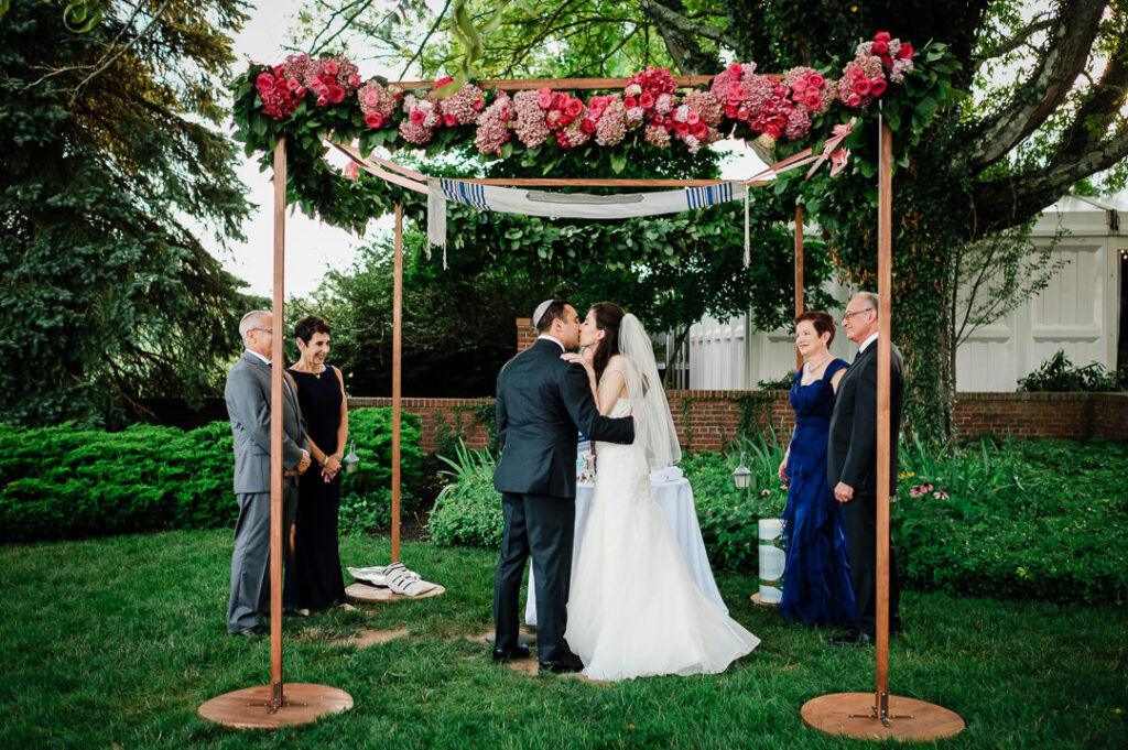 Newlyweds sharing first kiss under chuppah decorated with abundant pink and crimson flowers and greenery