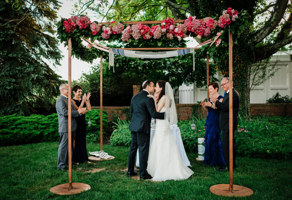 Parents applauding as couple shares first kiss under tallit-adorned chuppah at Hamilton Farm Jewish wedding