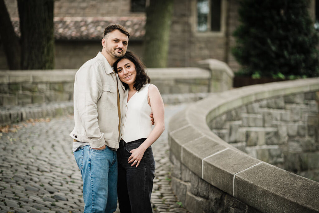 Casual engagement photo of couple walking on cobblestone path at Fort Tryon Park with Cloisters building in background