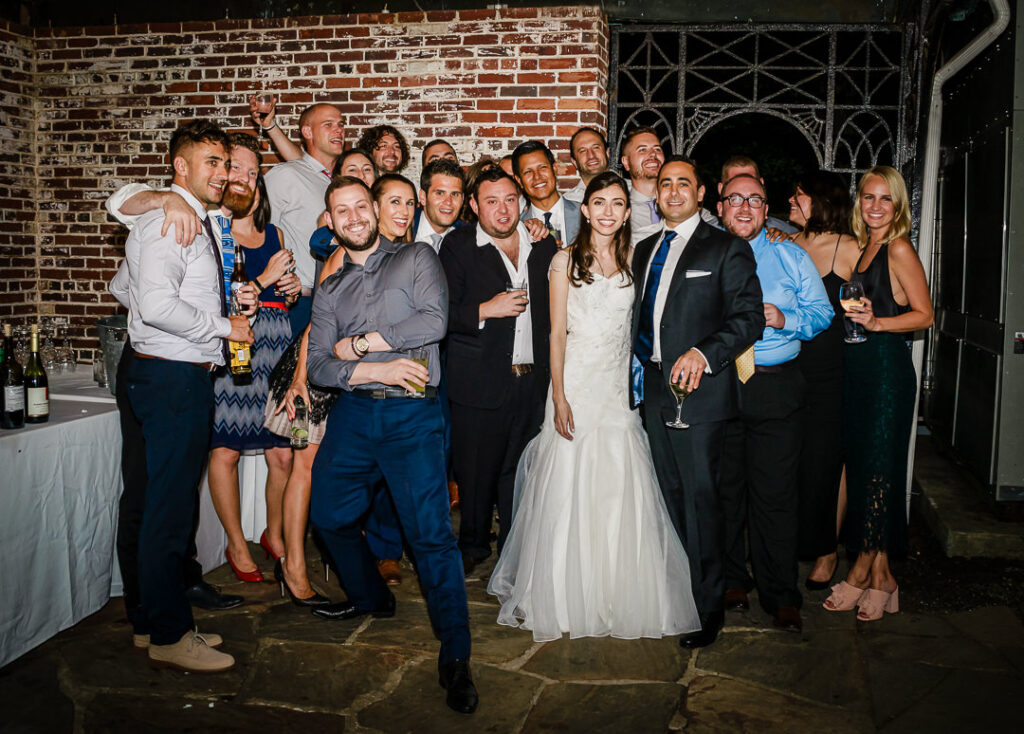 Large group of friends celebrating with bride and groom against exposed brick wall at late evening reception