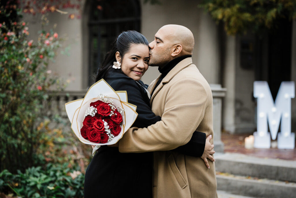 Groom kisses newly engaged bride on cheek as she holds red roses bouquet, romantic fall proposal photography Van Vleck Gardens Montclair NJ