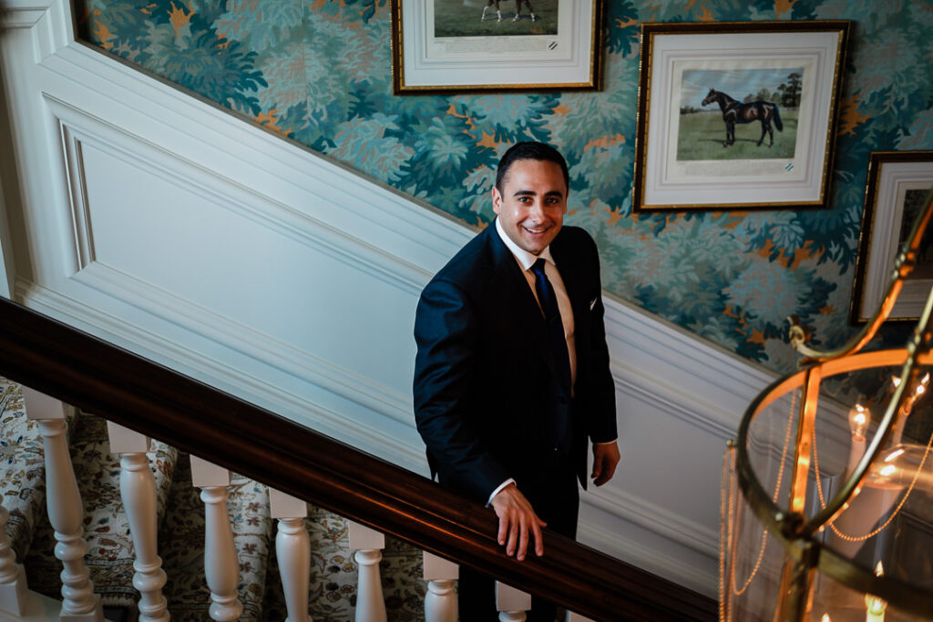 Groom in navy suit on elegant staircase with equestrian wallpaper at Hamilton Farm Golf Club wedding