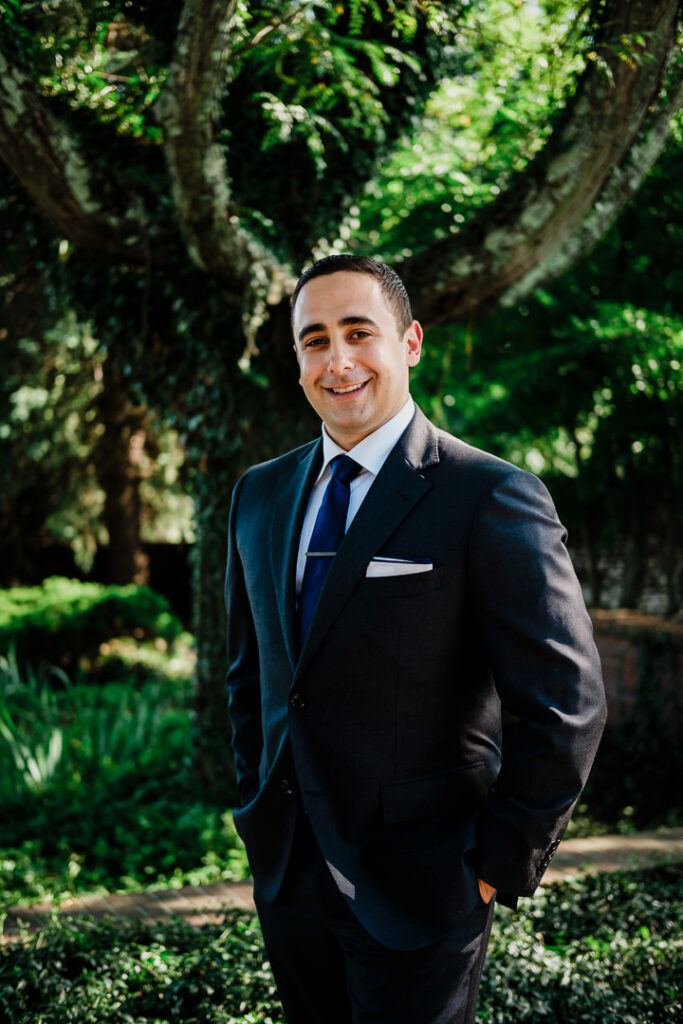 Handsome groom in navy suit and kippah smiling confidently under shade trees