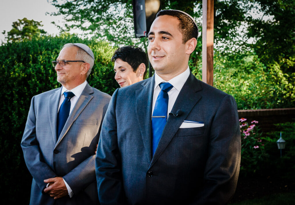 Emotional groom wearing kippah waiting under chuppah with parents before bride arrives at Jewish wedding