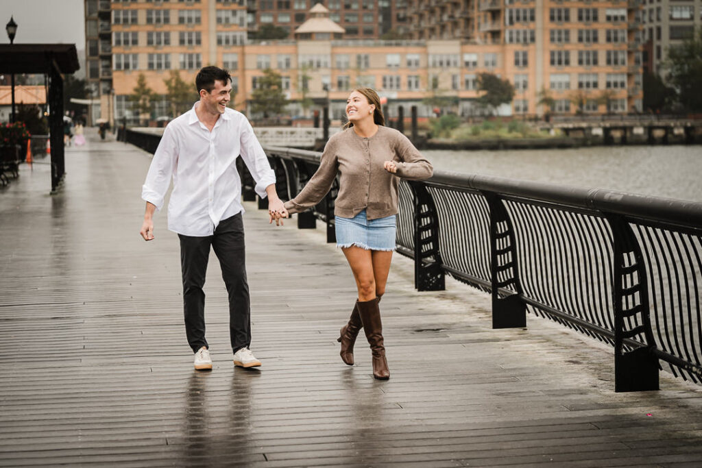 Newly engaged couple walking hand in hand on Pier 13 boardwalk after rainy proposal in Hoboken