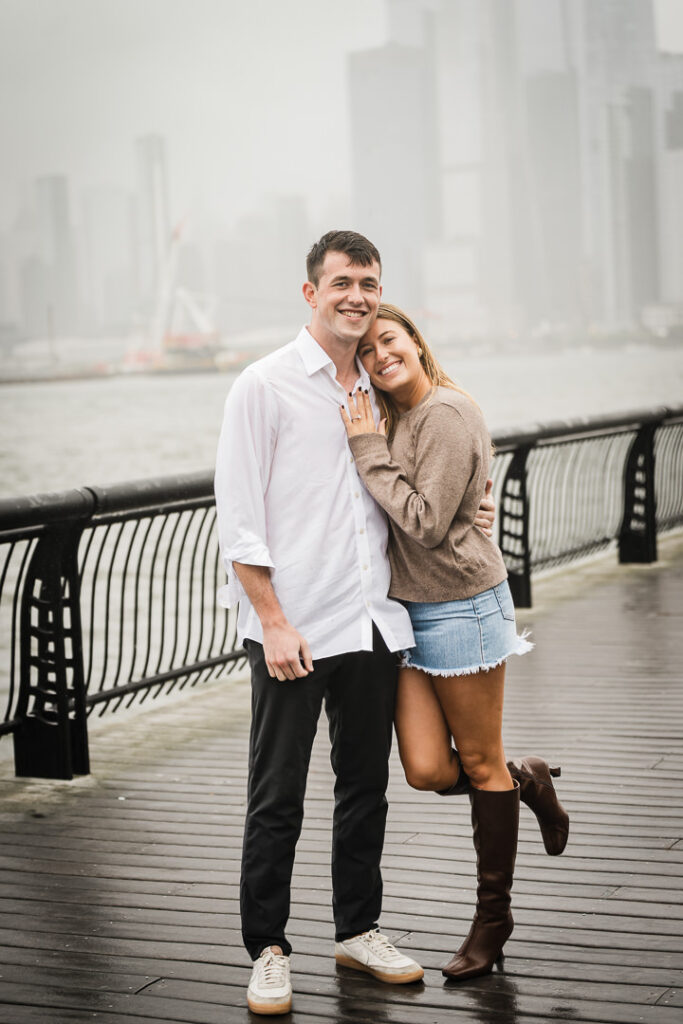 Joyful newly engaged couple leaning on each other at Pier 13 with rainy Hudson River view