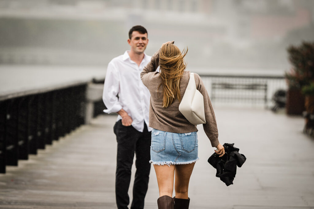 Couple walking on rainy Pier 13 boardwalk in Hoboken before surprise proposal with Manhattan skyline in misty background