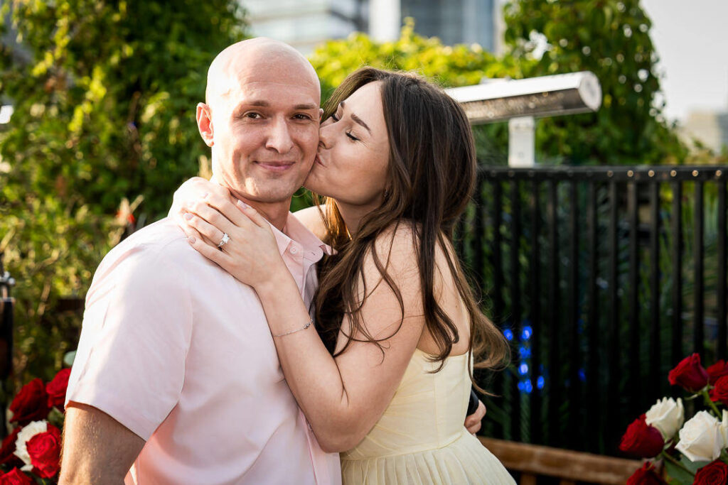 Intimate engagement portrait of bride kissing groom's cheek showing diamond ring, lush greenery and NYC skyline background