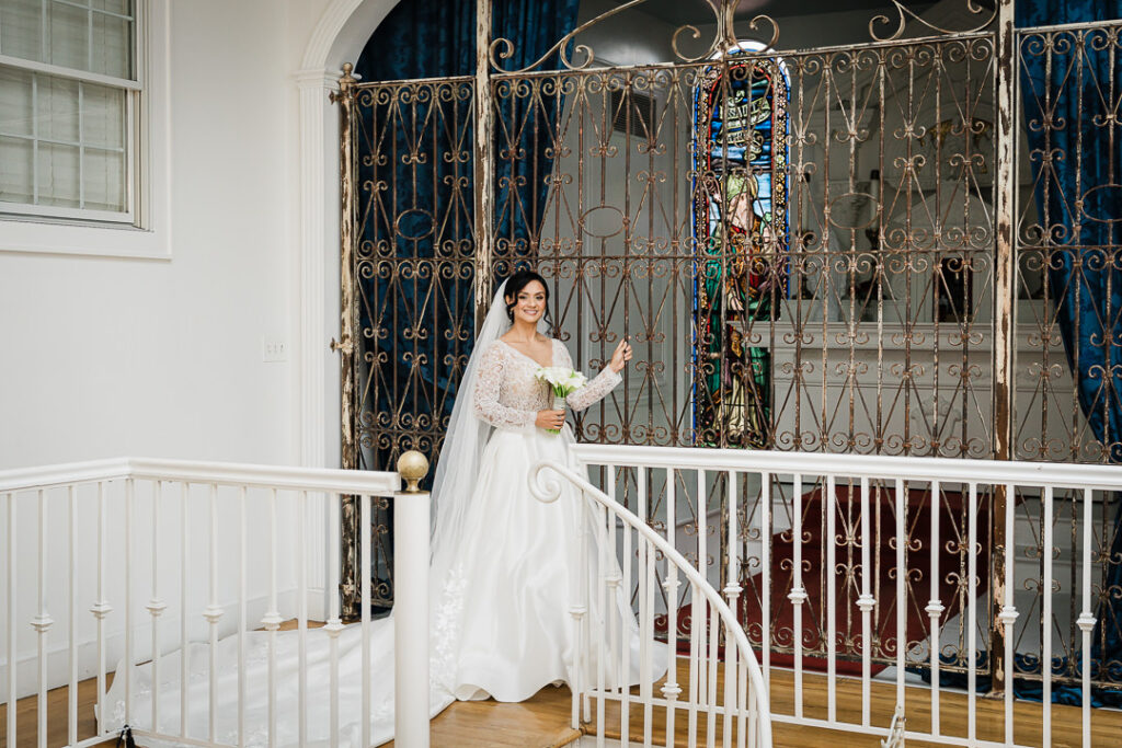 Bride with bouquet poses by ornate ironwork gate on staircase at James Ward Mansion Westfield wedding by Alex Kaplan