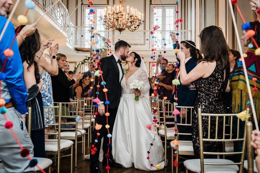 Newlyweds kissing during vibrant pom-pom garland ceremony exit James Ward Mansion by Alex Kaplan