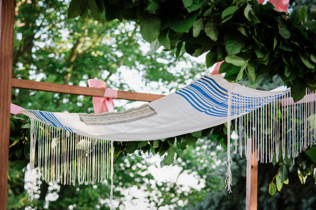 Traditional Jewish prayer shawl with blue stripes draped over wooden chuppah frame with pink ribbon accents