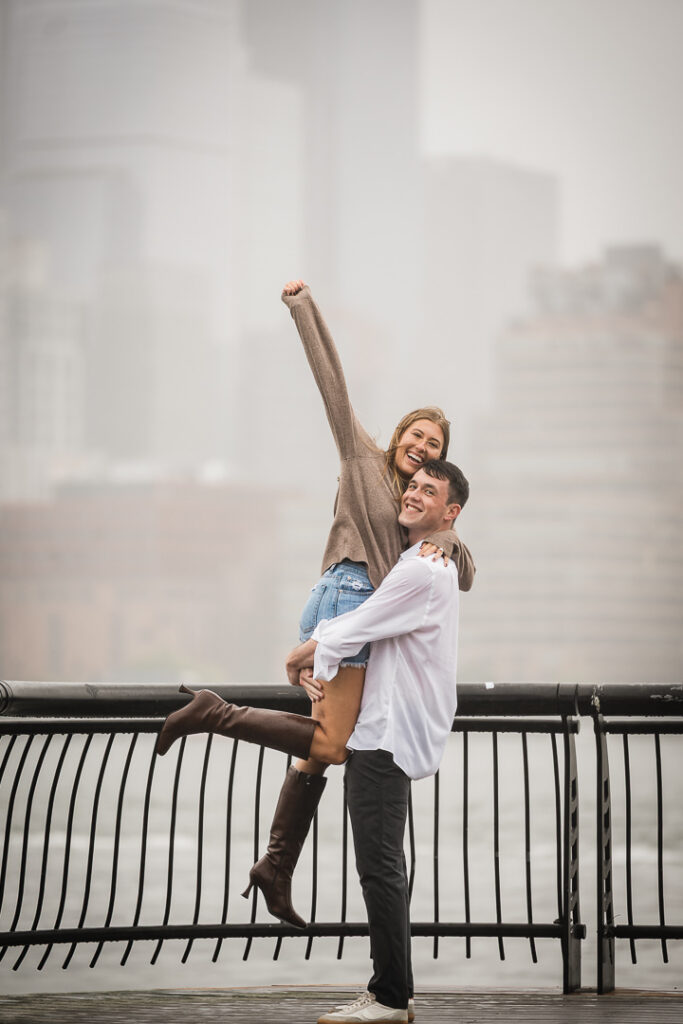 Excited newly engaged couple celebrating with arms raised on Pier 13 Hoboken during rainy proposal