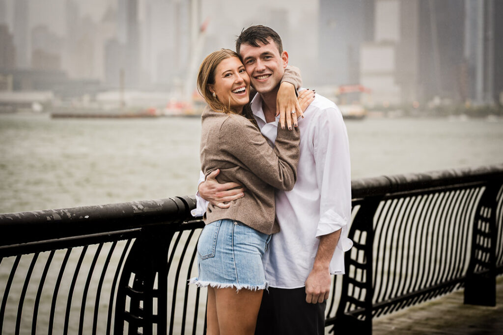 Happy newly engaged couple laughing together on Pier 13 waterfront with foggy river behind them