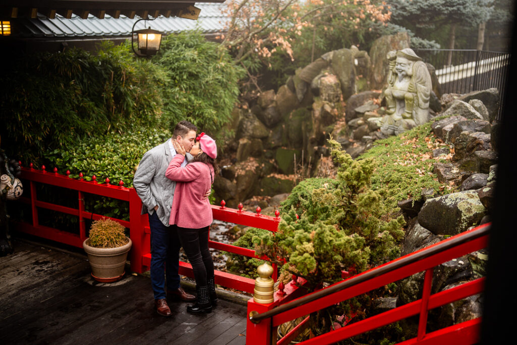 Engaged couple kissing on red garden bridge at The Views at Mount Fuji, NY.