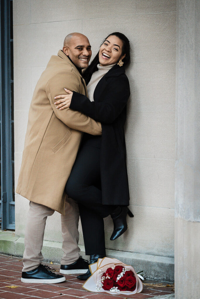 Engaged couple laughs together against Van Vleck garden wall with red roses bouquet, candid fall engagement photography Montclair NJ