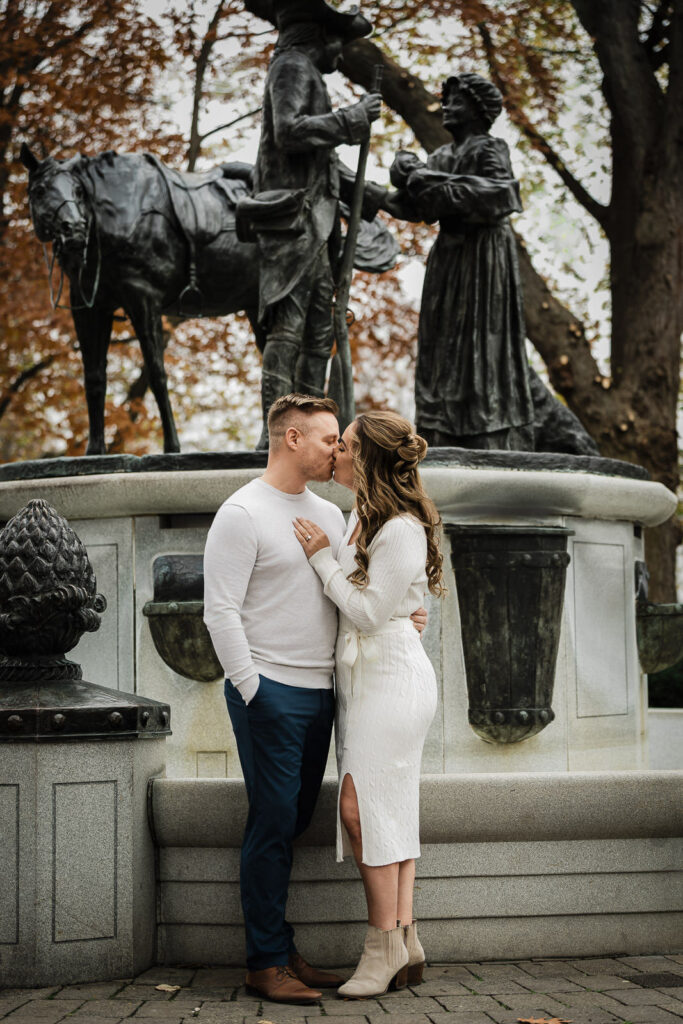 Engaged couple kissing at Morristown Green Civil War Monument during fall engagement session with historic bronze statue in background