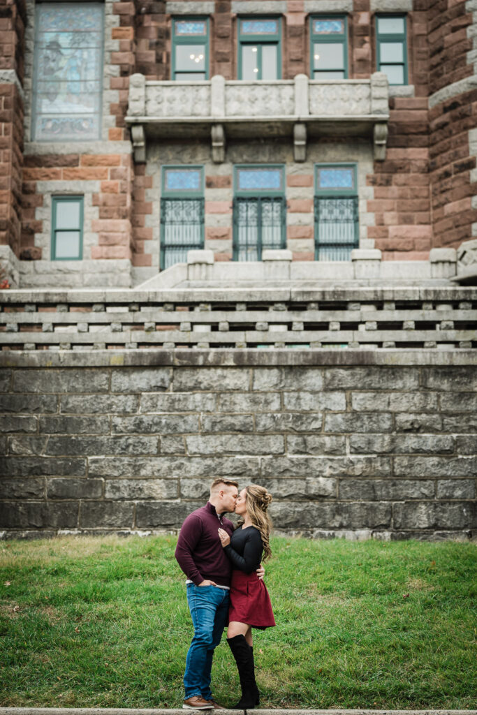 Couple kissing at historic Morristown building during engagement session wearing burgundy and black coordinated outfits
