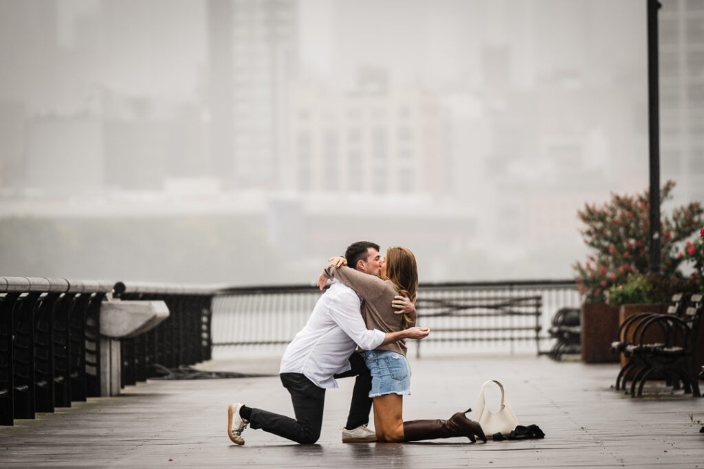 Newly engaged couple embracing and kissing on Pier 13 boardwalk after rainy Hoboken proposal