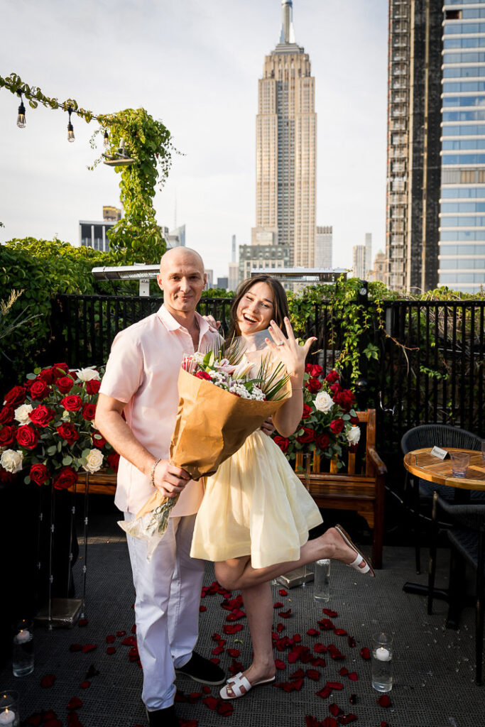 Happy newly engaged couple poses with flower bouquet and engagement ring, Empire State Building visible in background, NYC proposal photographer