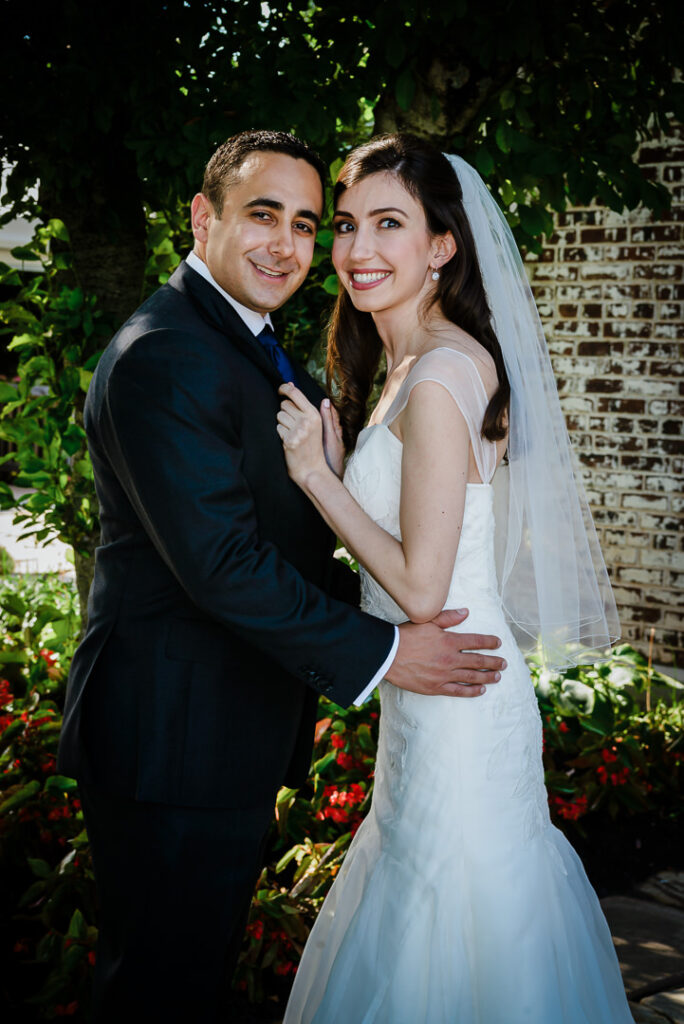 Newlywed couple in formal wedding attire surrounded by vibrant red begonias and mature trees