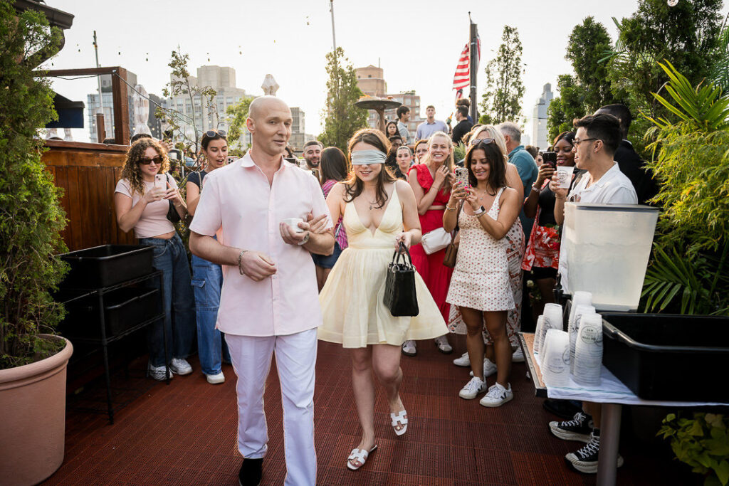 Surprised bride-to-be walks blindfolded through rooftop proposal setup with friends and family watching, NYC skyline and American flag visible, Northern NJ proposal photographer Alex Kaplan