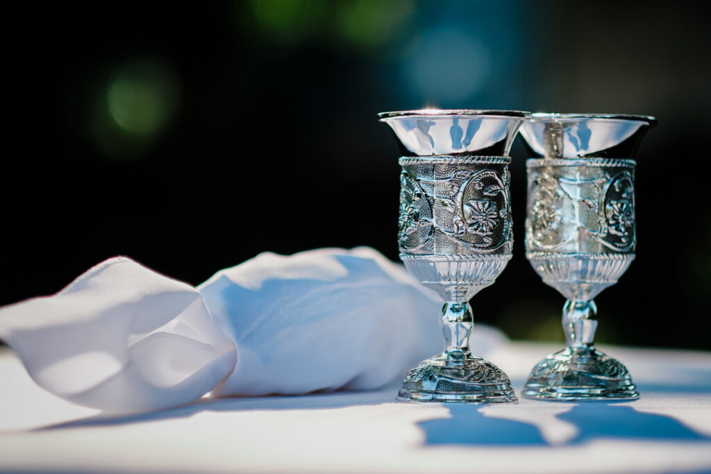 Detailed closeup of ornate silver Kiddush cups with intricate filigree designs and white tallit for Jewish wedding ceremony
