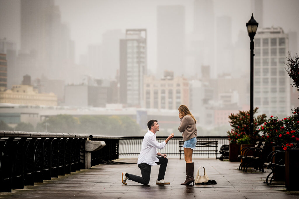 Man proposing on one knee to girlfriend at Pier 13 Hoboken with foggy Manhattan skyline behind them during rainy proposal