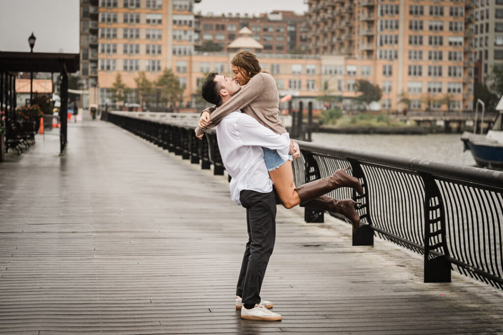 Groom lifting bride celebrating engagement at Pier 13 with Hoboken waterfront buildings in background