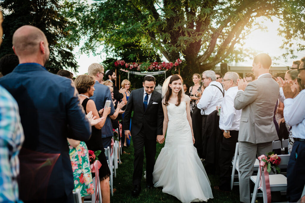 Joyful newlyweds walking back down aisle hand-in-hand with guests applauding during golden hour recessional