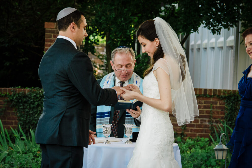 Couple exchanging rings under chuppah with rabbi wearing traditional tallit officiating Jewish wedding ceremony