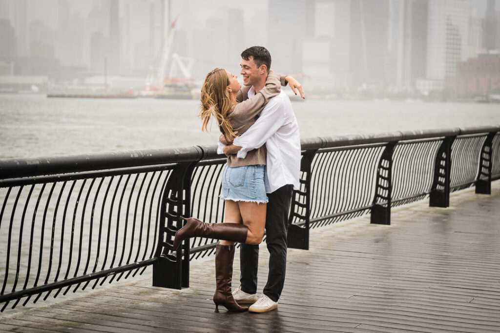Engaged couple in loving embrace on Pier 13 overlooking Hudson River with misty Manhattan in background