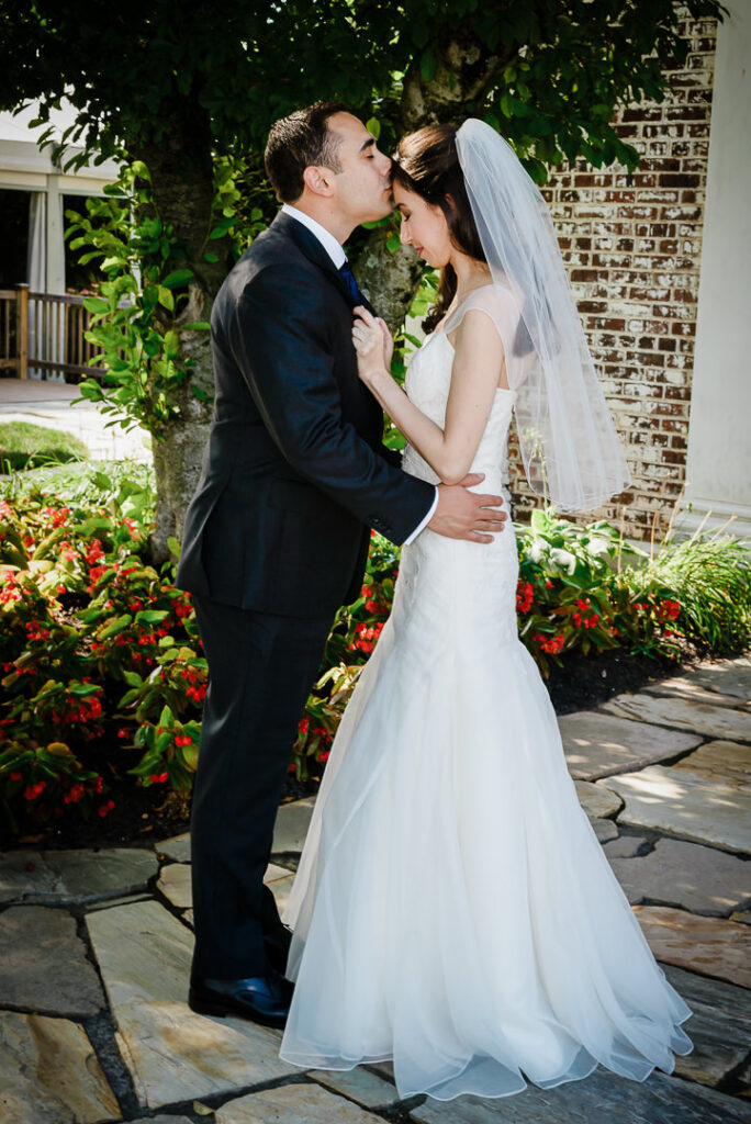 Groom kissing bride's forehead on flagstone pathway with colorful garden flowers at Hamilton Farm
