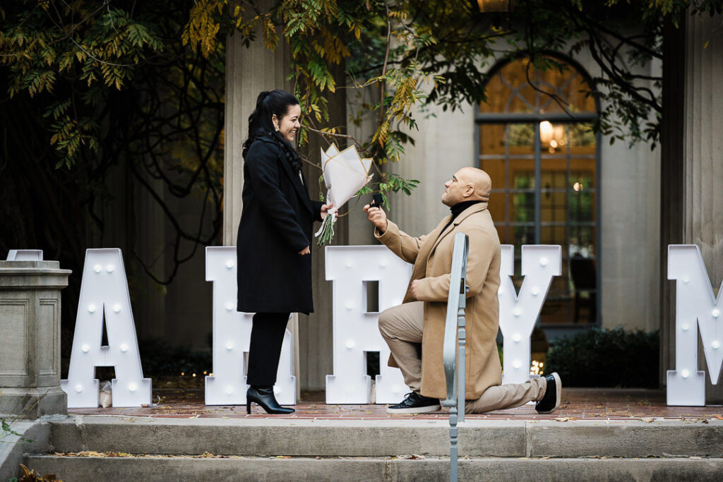 Groom proposes on one knee in front of illuminated MARRY ME letters at Van Vleck House & Gardens Montclair NJ, fall proposal photography by Alex Kaplan Northern New Jersey