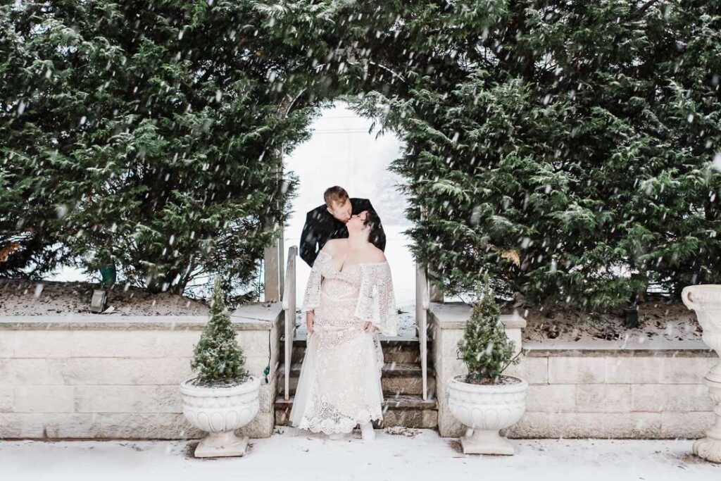 Bride and groom kissing under snowy evergreen arch during winter wedding captured by Alex Kaplan in New Jersey