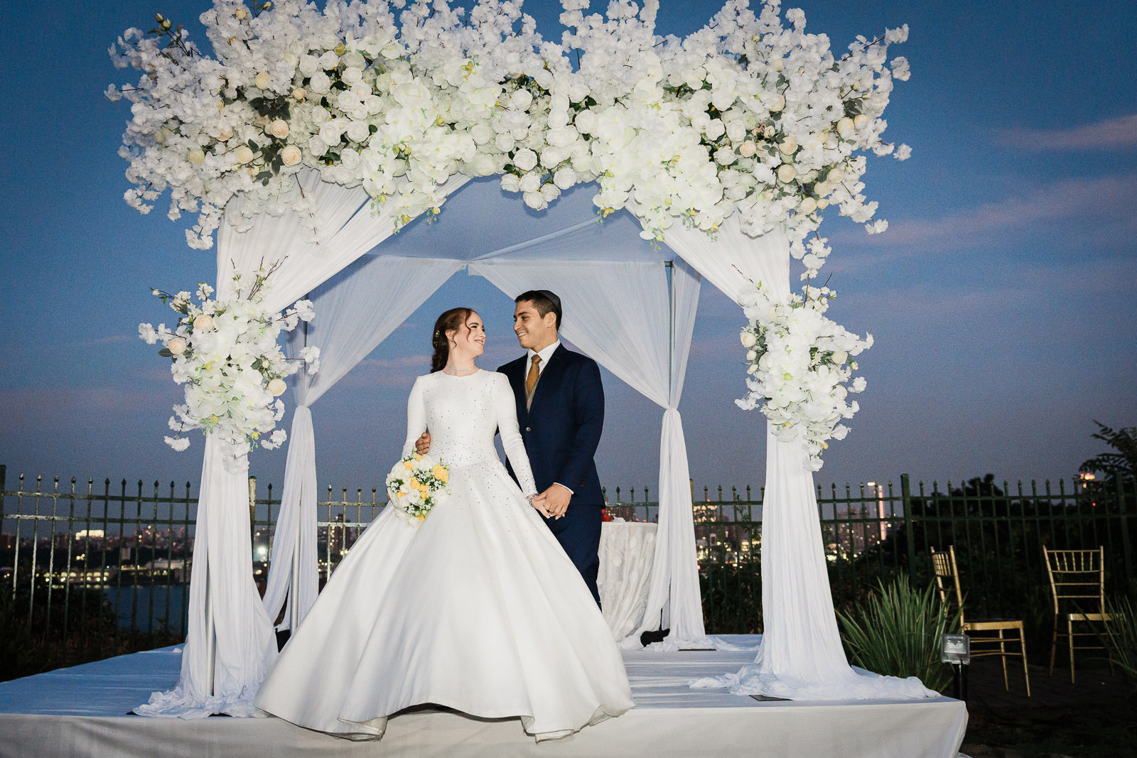 Bride and groom portrait under white floral chuppah at blue hour with NYC skyline at Riverview Ballroom Cliffside Park Orthodox Jewish wedding