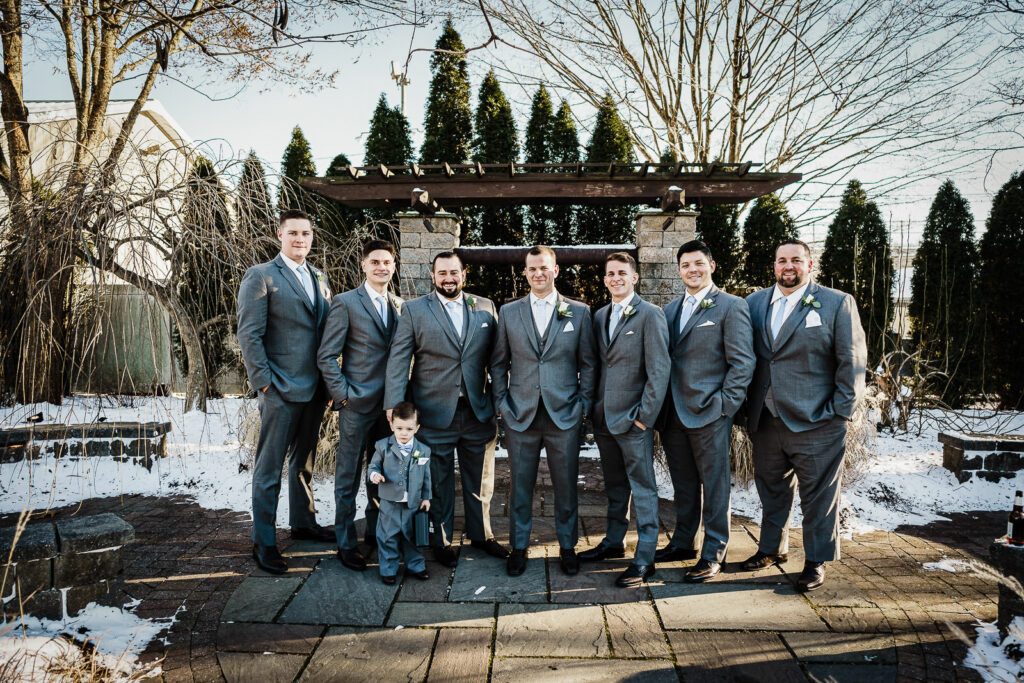 Groom and groomsmen in gray suits at snowy Sussex County Fairgrounds pergola photographed by Alex Kaplan