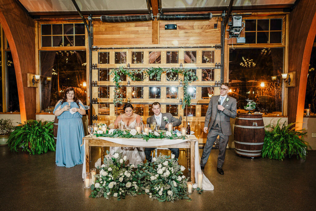 Maid of honor and best man giving toasts at sweetheart table with greenery backdrop at The Conservatory photographed by Alex Kaplan