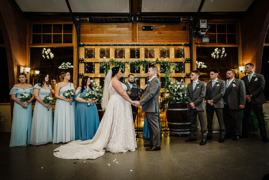 Indoor winter wedding ceremony with greenery arch and blue bridesmaids at The Conservatory at Sussex County Fairgrounds photographed by Alex Kaplan