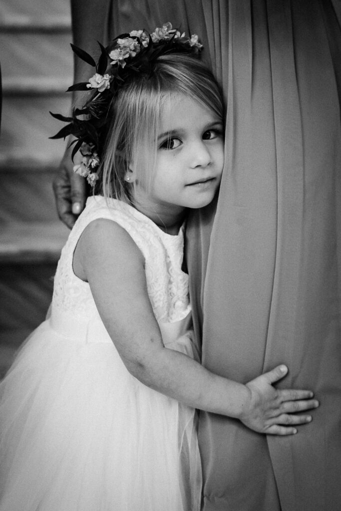 Adorable flower girl with floral crown peeking around curtain at Lucien's Manor wedding captured by NJ photographer Alex Kaplan