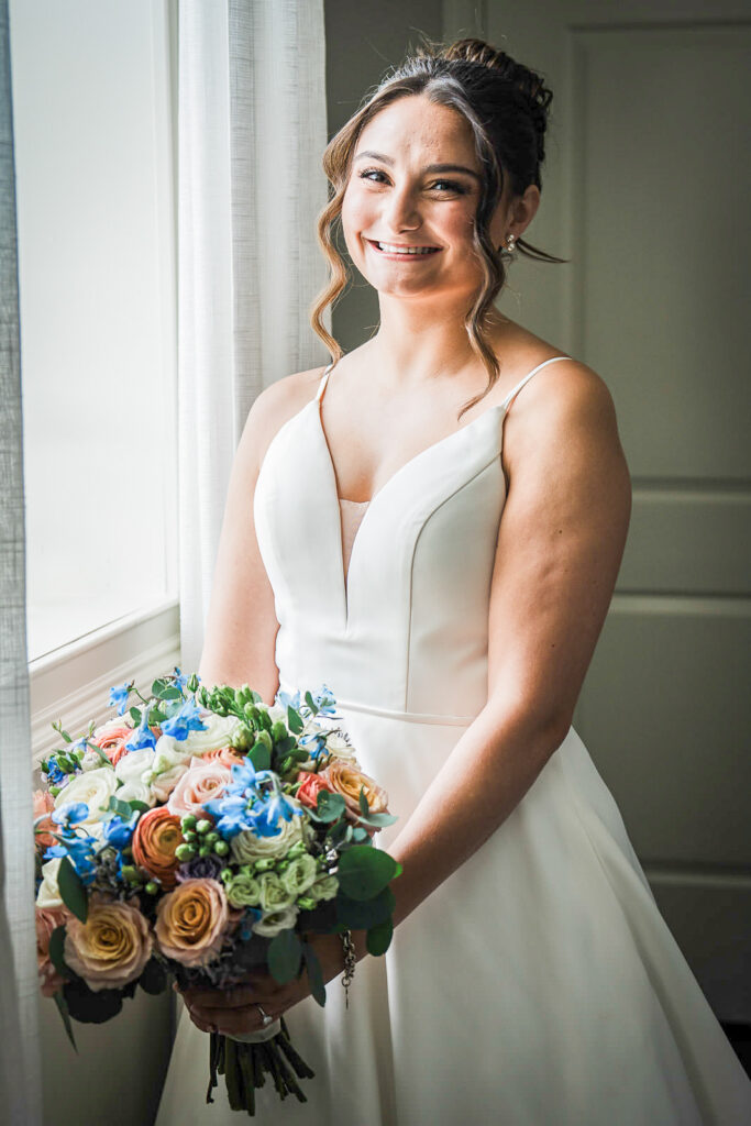 Bride holding colorful bouquet by window in natural light at Mansion at Mountain Lakes bridal suite