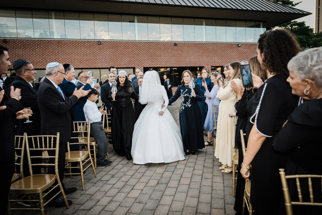 Bride walking down outdoor ceremony aisle with NYC skyline background at Riverview Ballroom Orthodox Jewish wedding