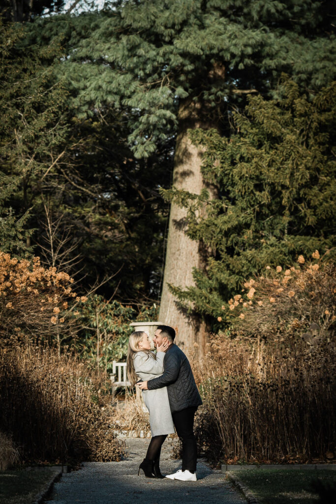 Engaged couple kissing under tall trees at Van Vleck House and Gardens Montclair New Jersey