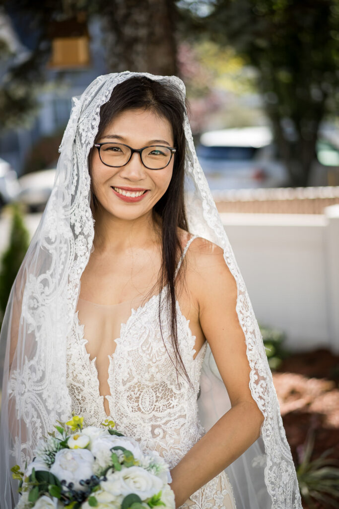 Bride Ruby in detailed lace wedding gown with cathedral veil smiling on Fair Lawn residential street holding white and green bouquet